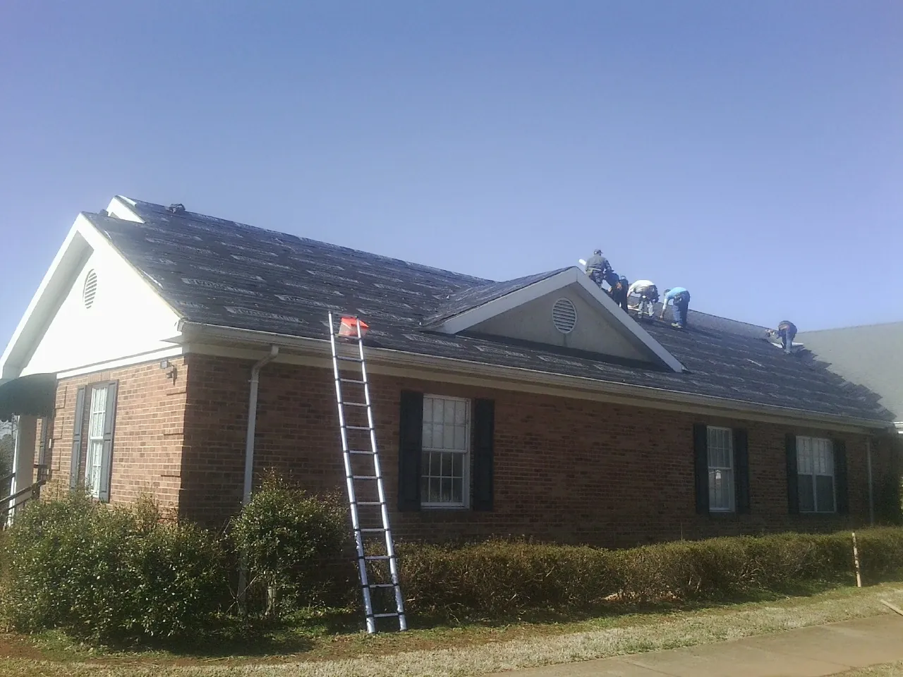 Crew tearing shingles off a brick ranch roof with a ladder set — Metal Roof Repair work by our crew in Otsego