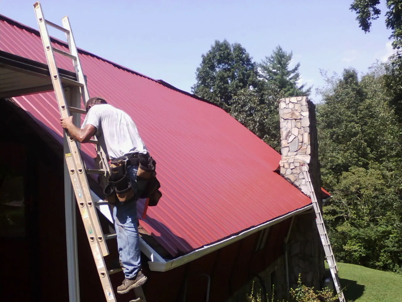Roofer on a ladder beside a red metal roof and stone chimney — Soffit & Fascia Repair by Roofing Brothers in Otsego